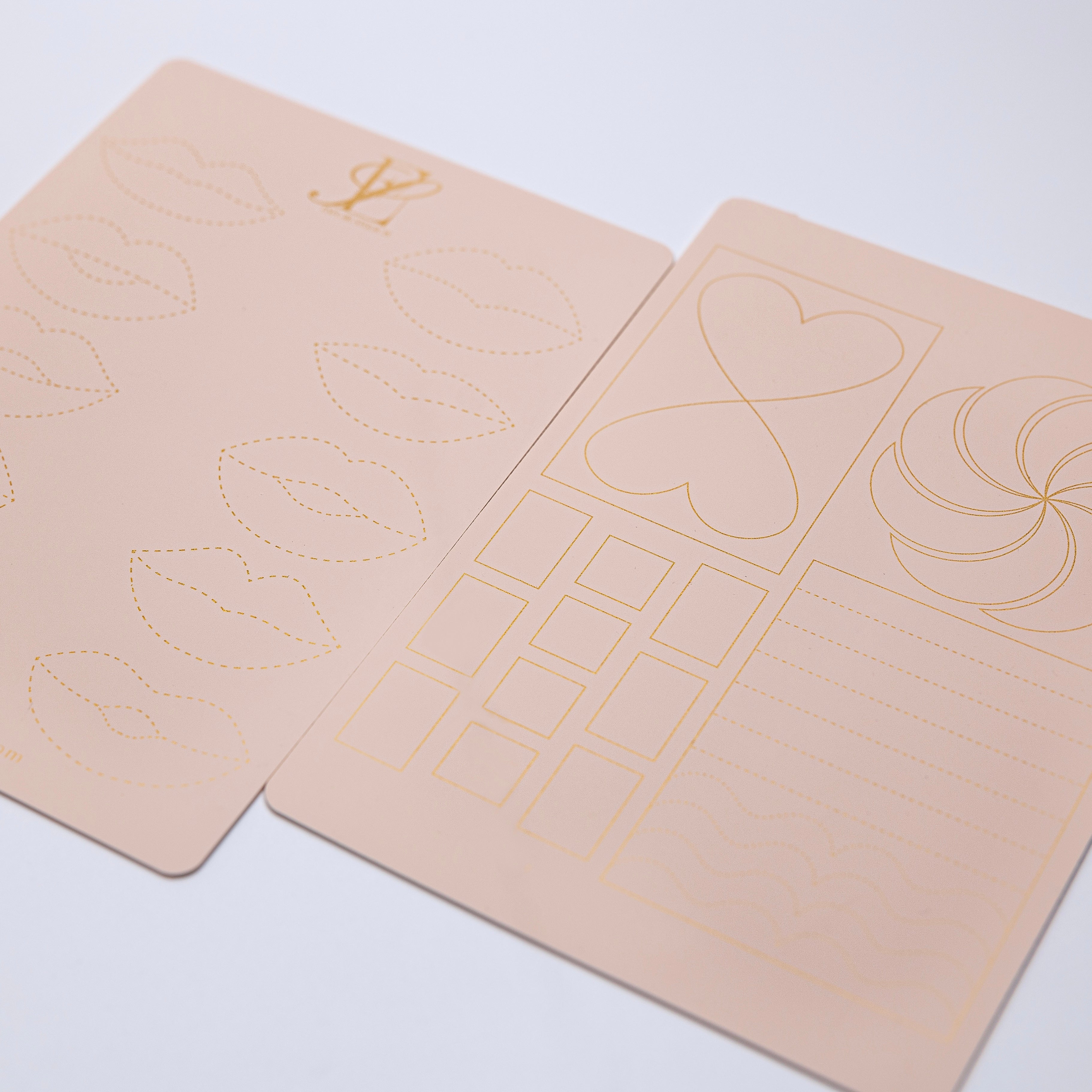 Two sheets of light pink paper with various golden line illustrations, including lips, hearts, squares, and wavy lines, arranged symmetrically on a light background. Perfect for enthusiasts of LIP PRACTICE PADS EXCLUSIVELY MADE BY ESPOIR STUDIOS by Espoir Studios.